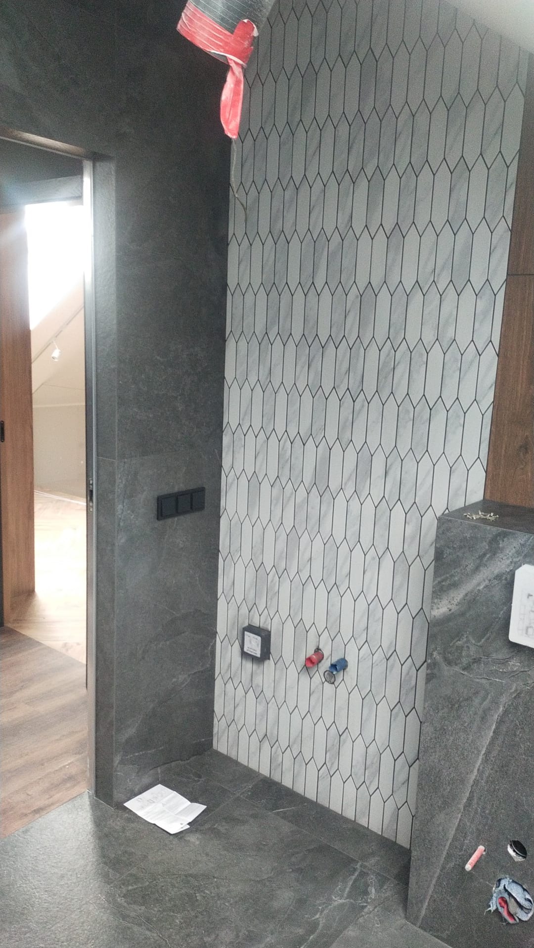 Wnętrze łazienki w trakcie remontu: położona szara mozaika na ścianie, wystające rury do podłączenia baterii, ciemna podłoga z płytek imitujących kamień, widoczny fragment instrukcji na podłodze...