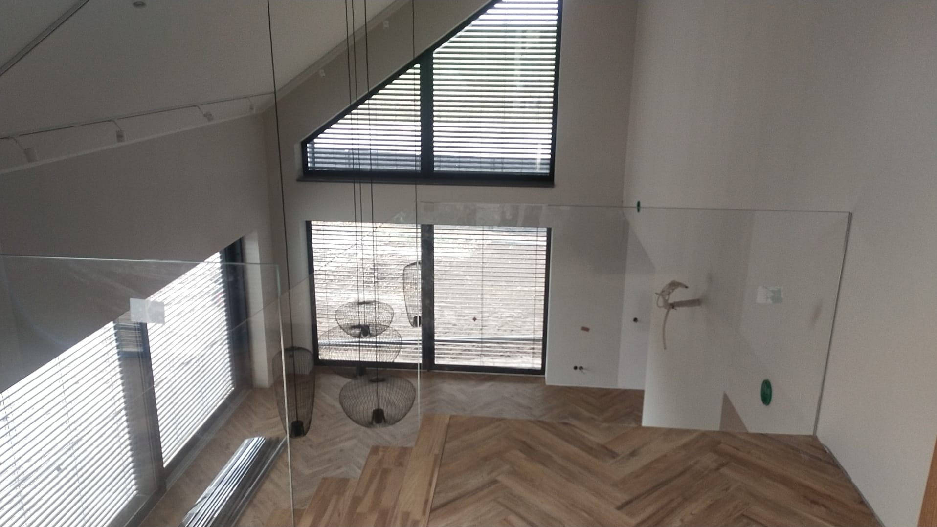 Wnętrze domu w stanie deweloperskim z widoczną drewnianą podłogą ułożoną w jodełkę, szklaną balustradą schodów, dużymi oknami z żaluzjami i wiszącymi lampami drucianymi.