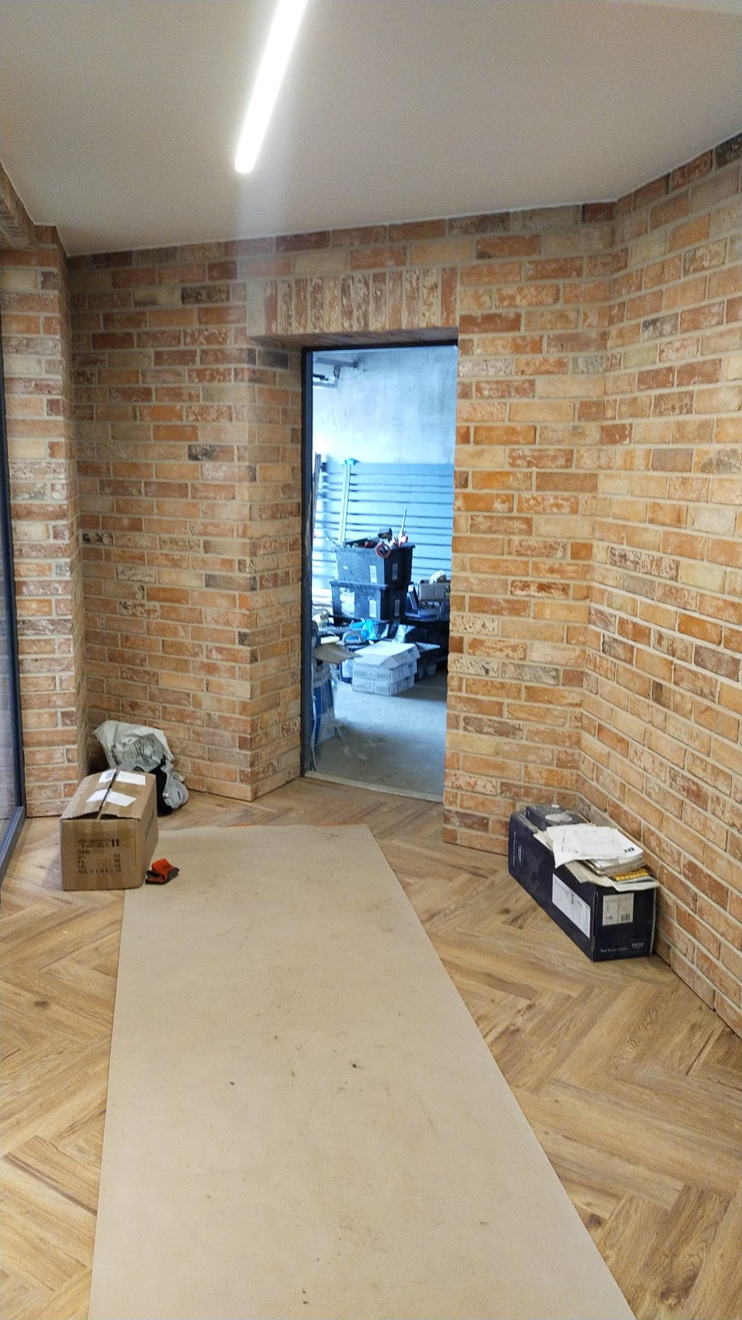 Wnętrze w trakcie wykańczania z ceglaną ścianą, widoczne kartony, płyta ochronna na podłodze i otwarte przejście do pomieszczenia z regałami.