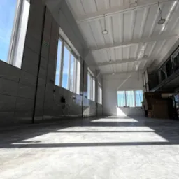 Jasna, przestronna hala przemysłowa z betonową posadzką, widoczne okna z metalowymi kratami i szare płytki na ścianach.