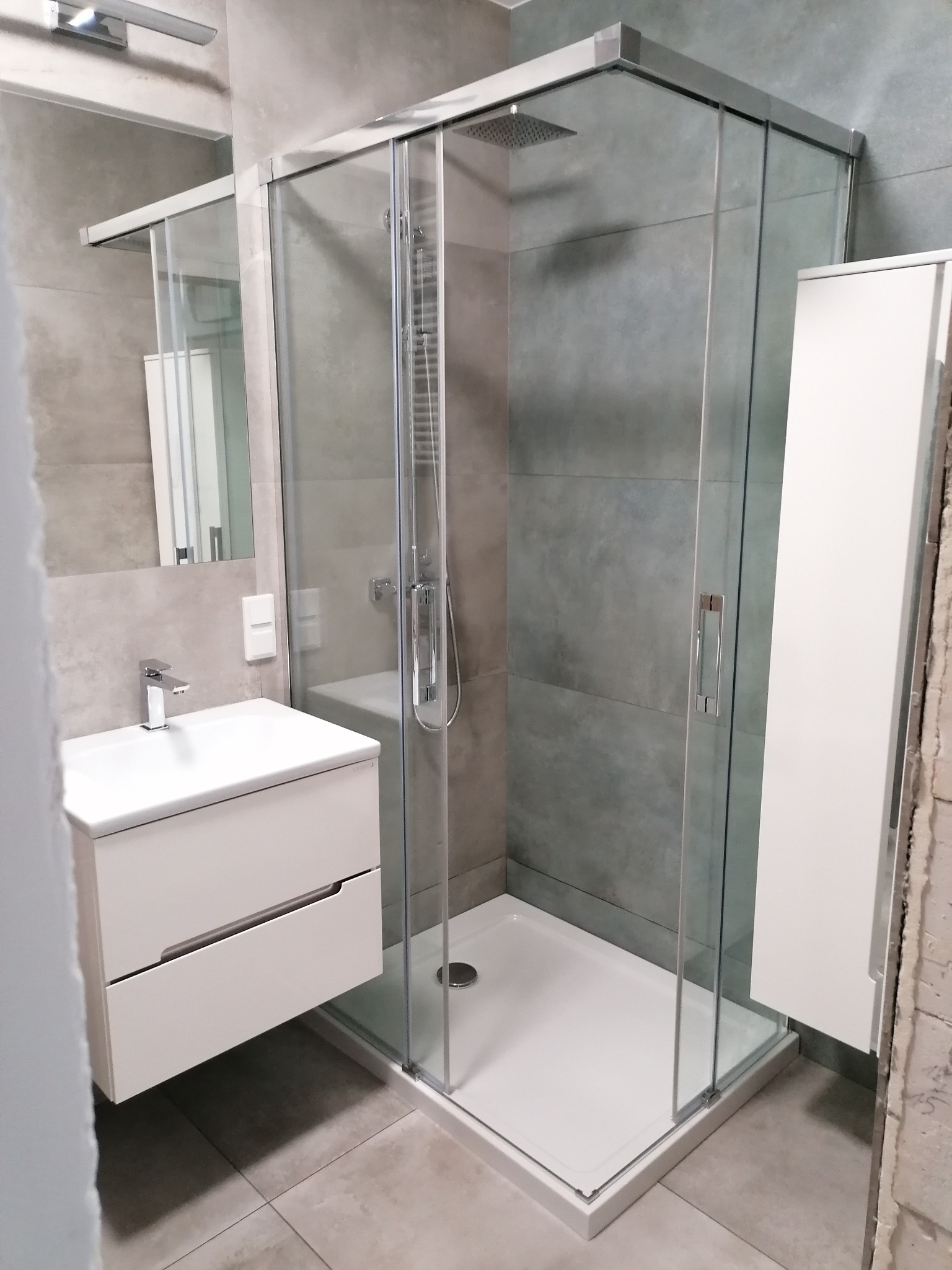 Nowoczesna łazienka z kabiną prysznicową typu walk-in, umywalką z szafką i szarą glazurą. Fragment ściany w stanie surowym dodaje surowego charakteru.