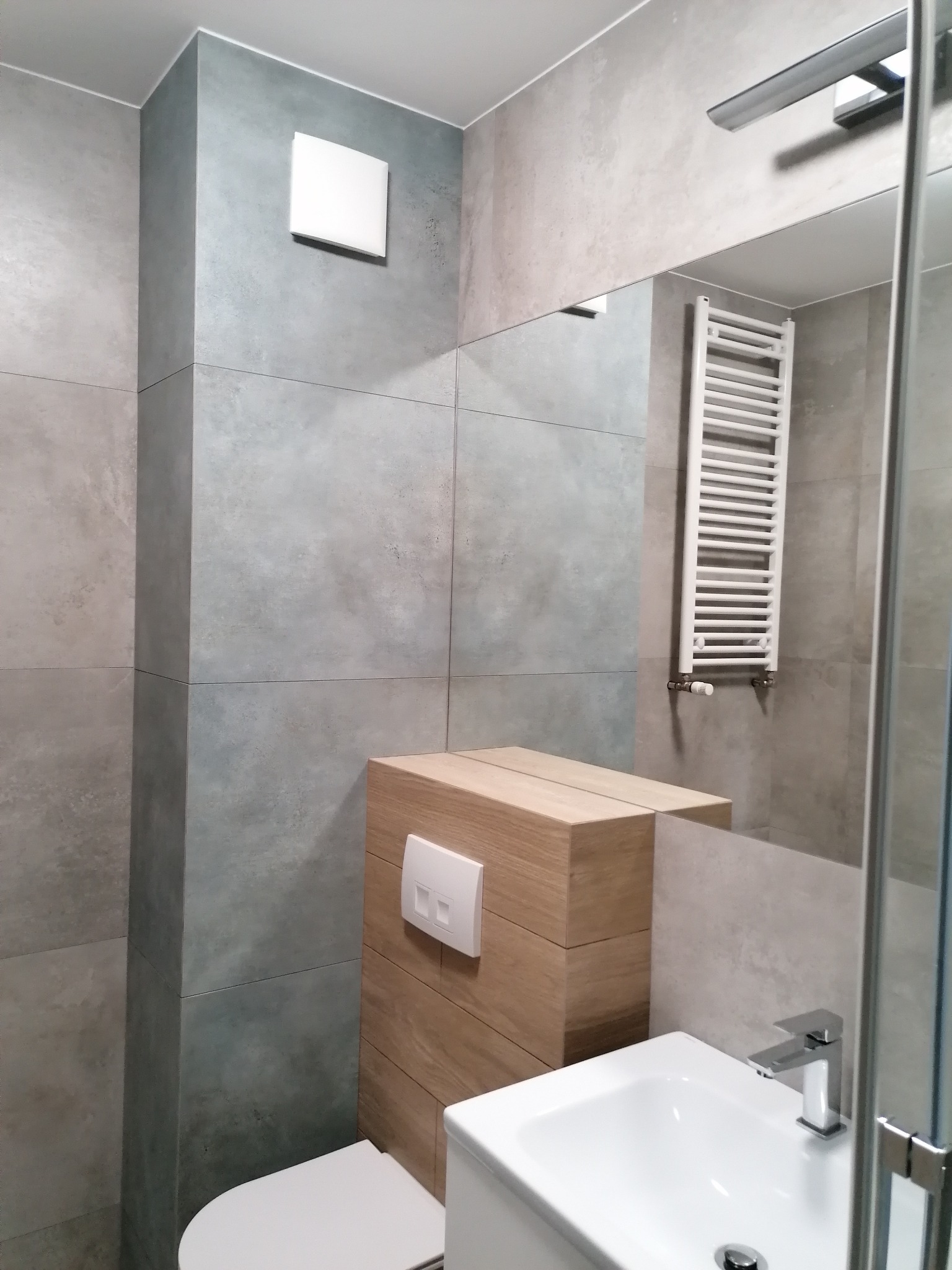 Nowoczesna łazienka z szarymi płytkami, drewnianą zabudową toalety i białą umywalką. W lustrze odbija się grzejnik drabinkowy. Minimalistyczny design.