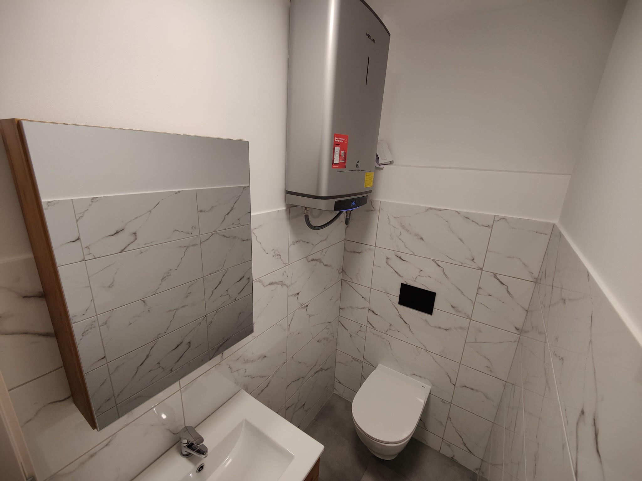 Wykończona łazienka z marmurowymi płytkami, wiszącą toaletą i grzejnikiem elektrycznym. Nowoczesny design, jasne kolory, minimalistyczny styl.