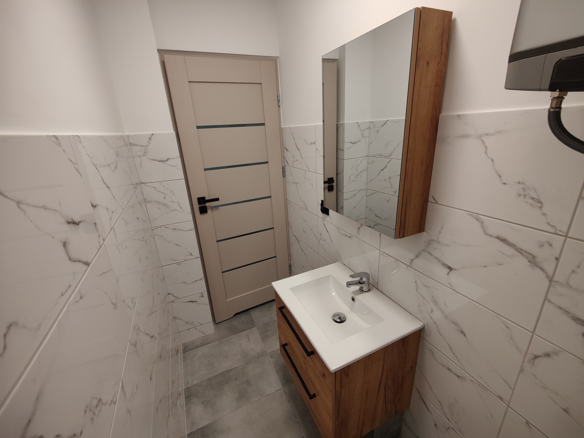 Nowoczesna łazienka z marmurowymi płytkami, drewnianą szafką pod umywalką i lustrem. Minimalistyczny design, jasne kolory, widoczny fragment bojlera.