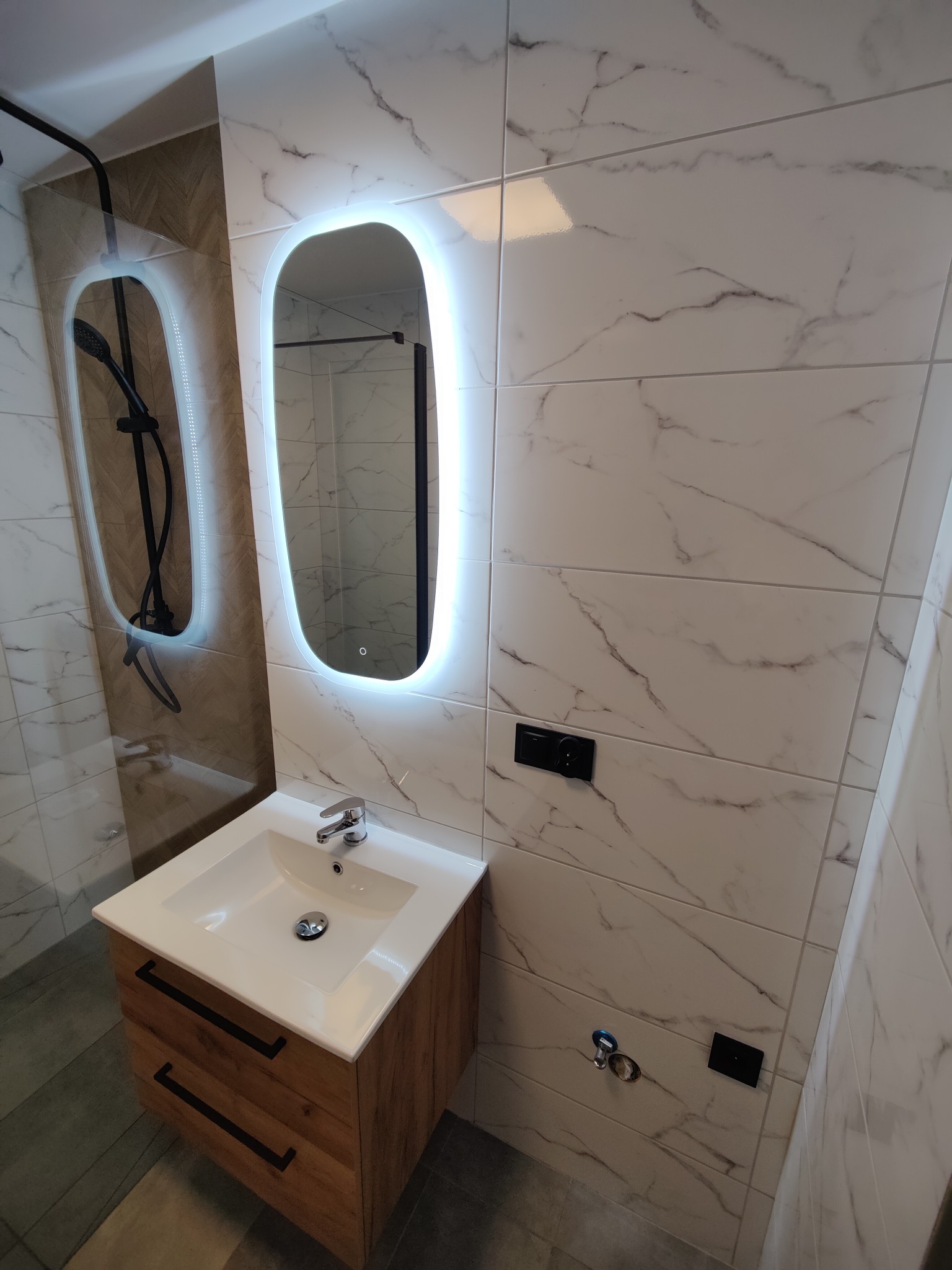 Nowoczesna łazienka z umywalką na szafce z drewnianym frontem, podświetlanym lustrem i marmurowymi płytkami na ścianach. Widoczna kabina prysznicowa.