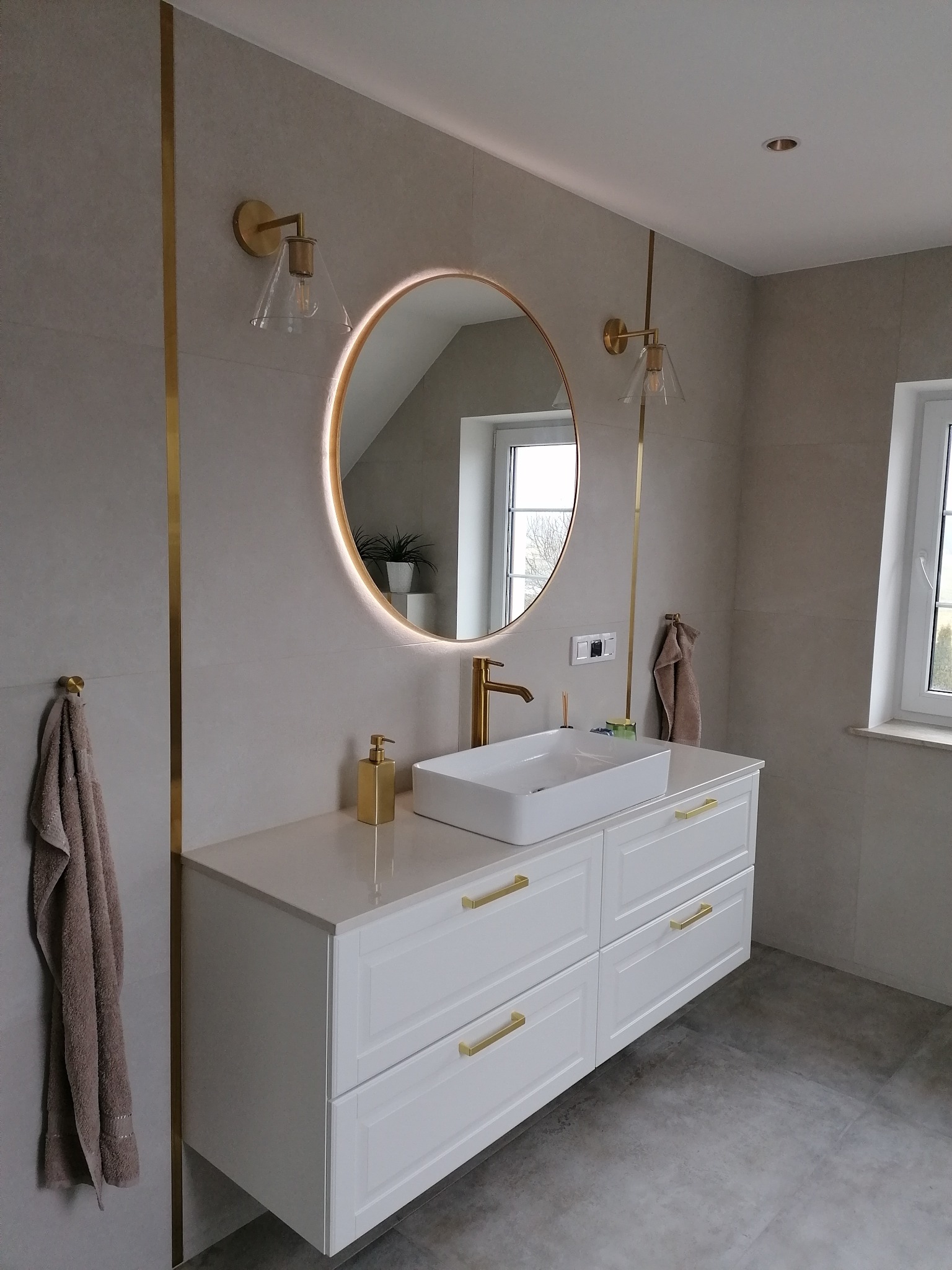 Elegancka łazienka z podświetlanym lustrem, białą szafką pod umywalką i złotymi akcentami. Widoczne płytki w odcieniach szarości i złote listwy dekoracyjne.