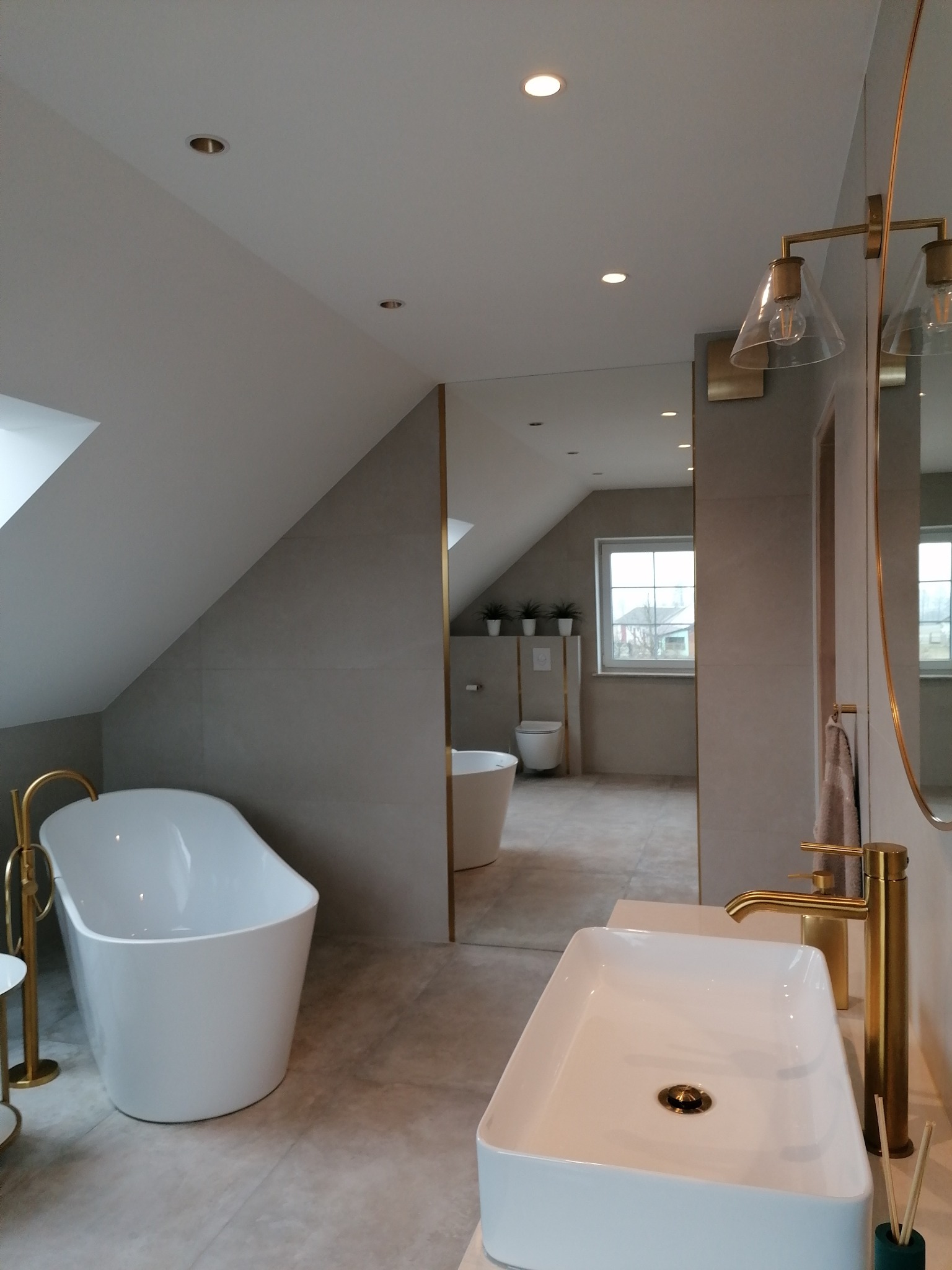 Nowoczesna łazienka na poddaszu z wolnostojącą wanną, umywalką nablatową i złotą armaturą. Widok przez lustro na toaletę i okno dachowe.
