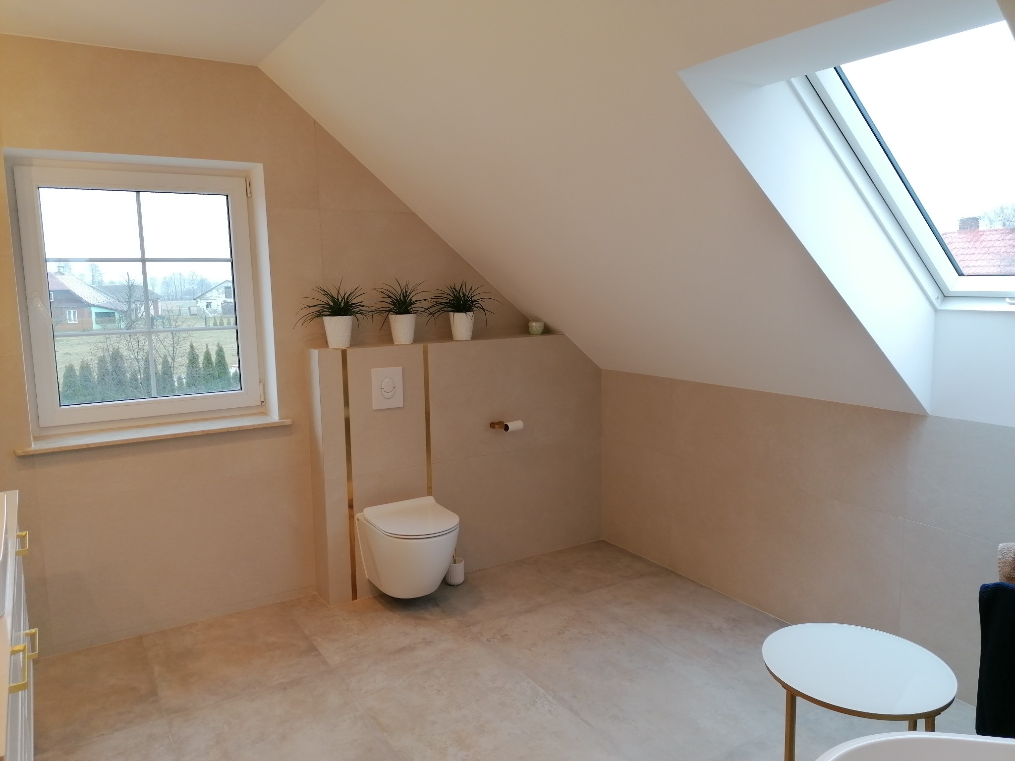 Jasna łazienka na poddaszu ze skośnym sufitem i oknem dachowym. Uwagę przykuwa podwieszana toaleta z subtelnymi złotymi akcentami i rośliny w doniczkach.