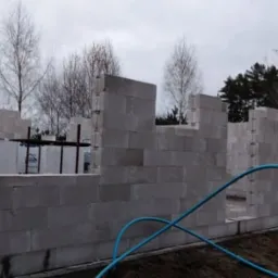 Budowa domu z betonu komórkowego: surowy stan ścian z widocznymi otworami okiennymi i drzwiowymi, na tle drzew i pochmurnego nieba, z niebieskim wężem ogrodowym na pierwszym planie.