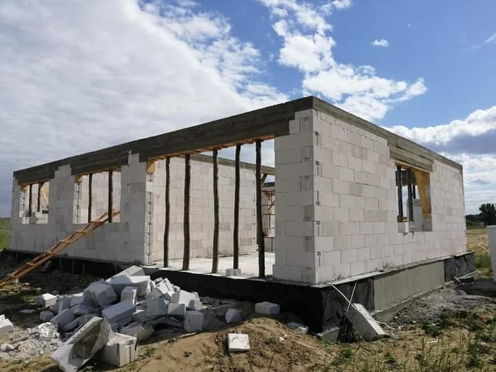 Budowa domu jednorodzinnego z białych bloczków w Toruniu, widoczne ściany w stanie surowym otwartym, drewniane stemple podtrzymujące strop, stos bloczków na ziemi, drabina oparta o ścianę.