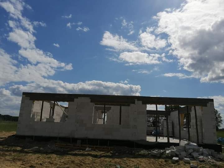 Surowy stan domu w trakcie budowy z bloczków, widoczne drewniane belki stropowe i niebo z chmurami w tle.