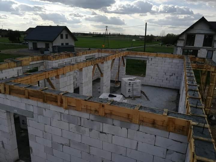 Wznoszenie ścian domu murowanego z betonu komórkowego, widoczne drewniane szalunki i zalany beton, w tle krajobraz z dwoma domami i polami pod pochmurnym niebem.