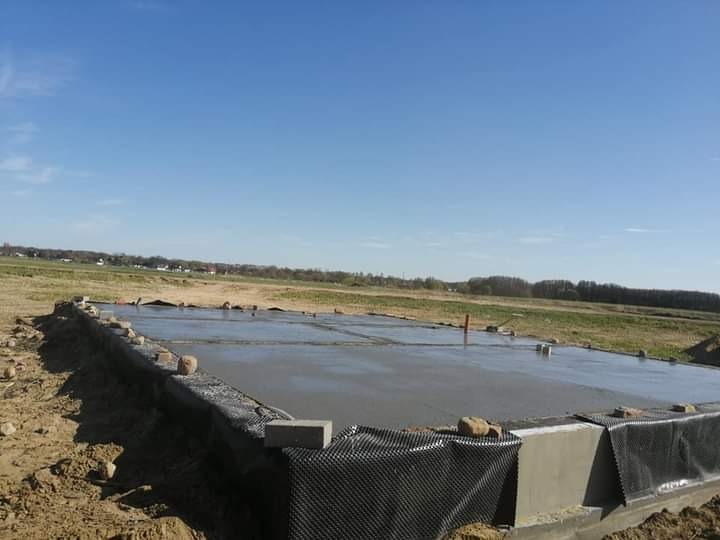 Świeżo wylana, gładka płyta fundamentowa pod budynek, zabezpieczona czarną folią drenażową, widok na pole i błękitne niebo.