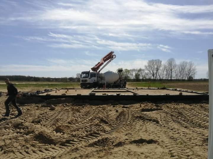 Betoniarka z podnośnikiem w trakcie wylewania fundamentów pod budynek na piaszczystym terenie, widoczny pracownik i obrys fundamentów.