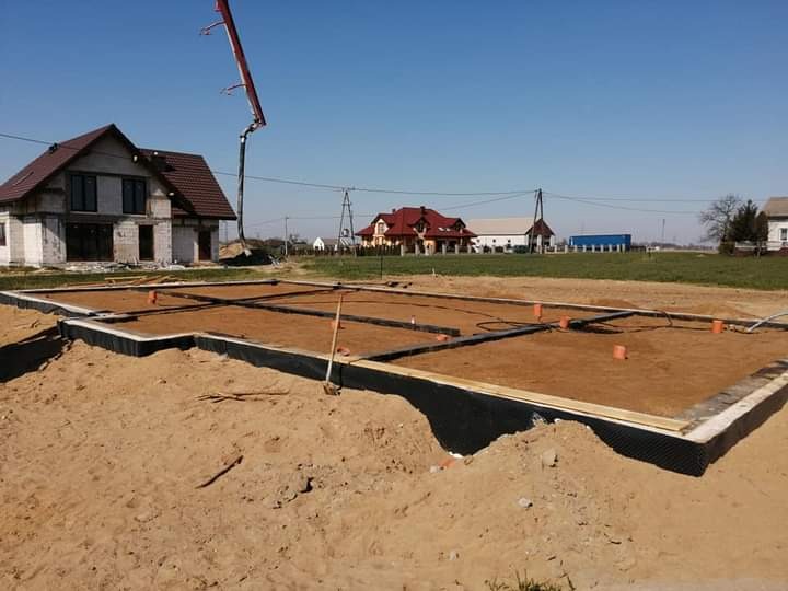 Fundamenty domu murowanego w trakcie budowy, widoczna izolacja przeciwwilgociowa, piasek i częściowo wzniesione ściany budynku w tle, ramię pompy do betonu w górnej części kadru.