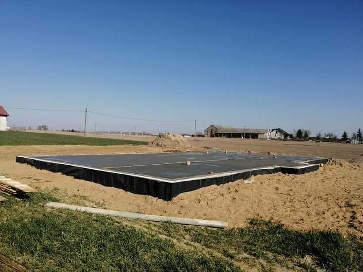 Wylany fundament pod dom jednorodzinny z izolacją pionową, widok na pole i budynki gospodarcze w tle.