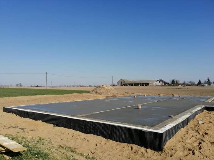 Wylany fundament pod budowę domu jednorodzinnego z izolacją pionową, widok na pole i budynki gospodarcze w tle, słoneczny dzień.