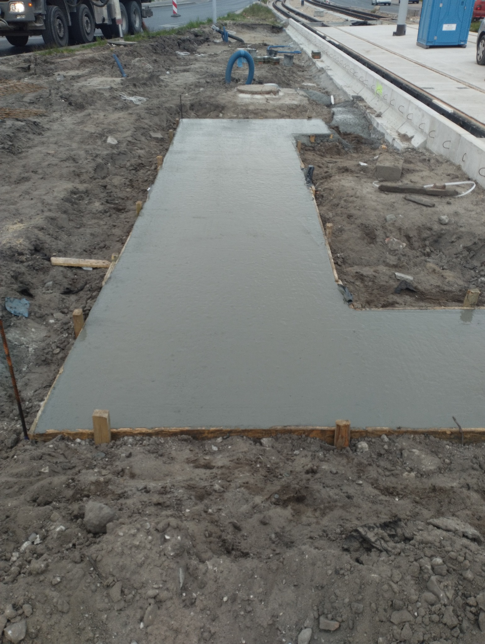 Świeżo wylana, mokra, betonowa ścieżka w szalunku z drewnianych desek na placu budowy, obok torów tramwajowych i przenośnej toalety.