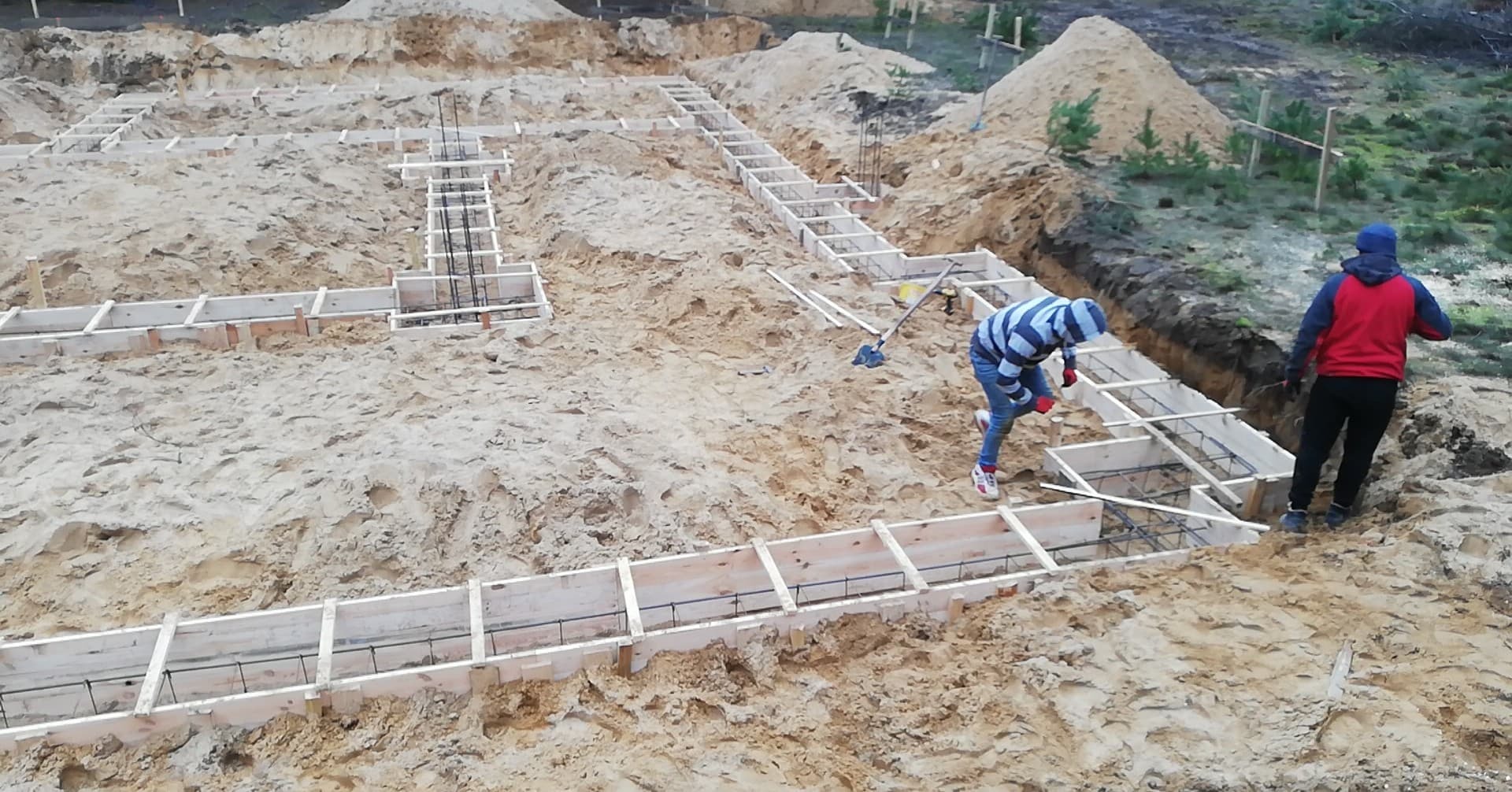Przygotowanie szalunków i zbrojenia pod fundament domu murowanego na piaszczystym terenie, widoczni dwaj pracownicy w trakcie pracy.
