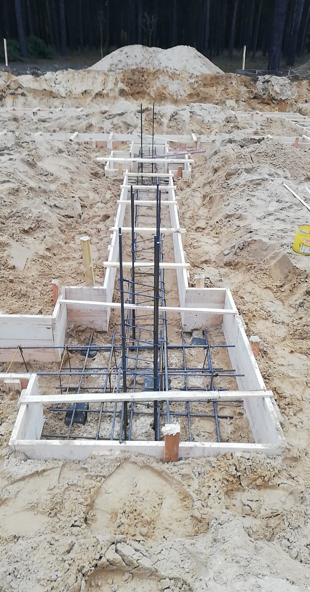 Przygotowanie fundamentów pod budowę: szalunki z desek, zbrojenie stalowe, wykop w piasku, widok z góry na plac budowy.