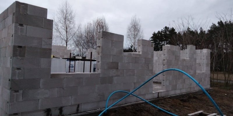 Budowa domu z betonu komórkowego: surowy stan ścian z widocznymi otworami okiennymi i drzwiowymi, na tle drzew i pochmurnego nieba, z niebieskim wężem ogrodowym na pierwszym planie.