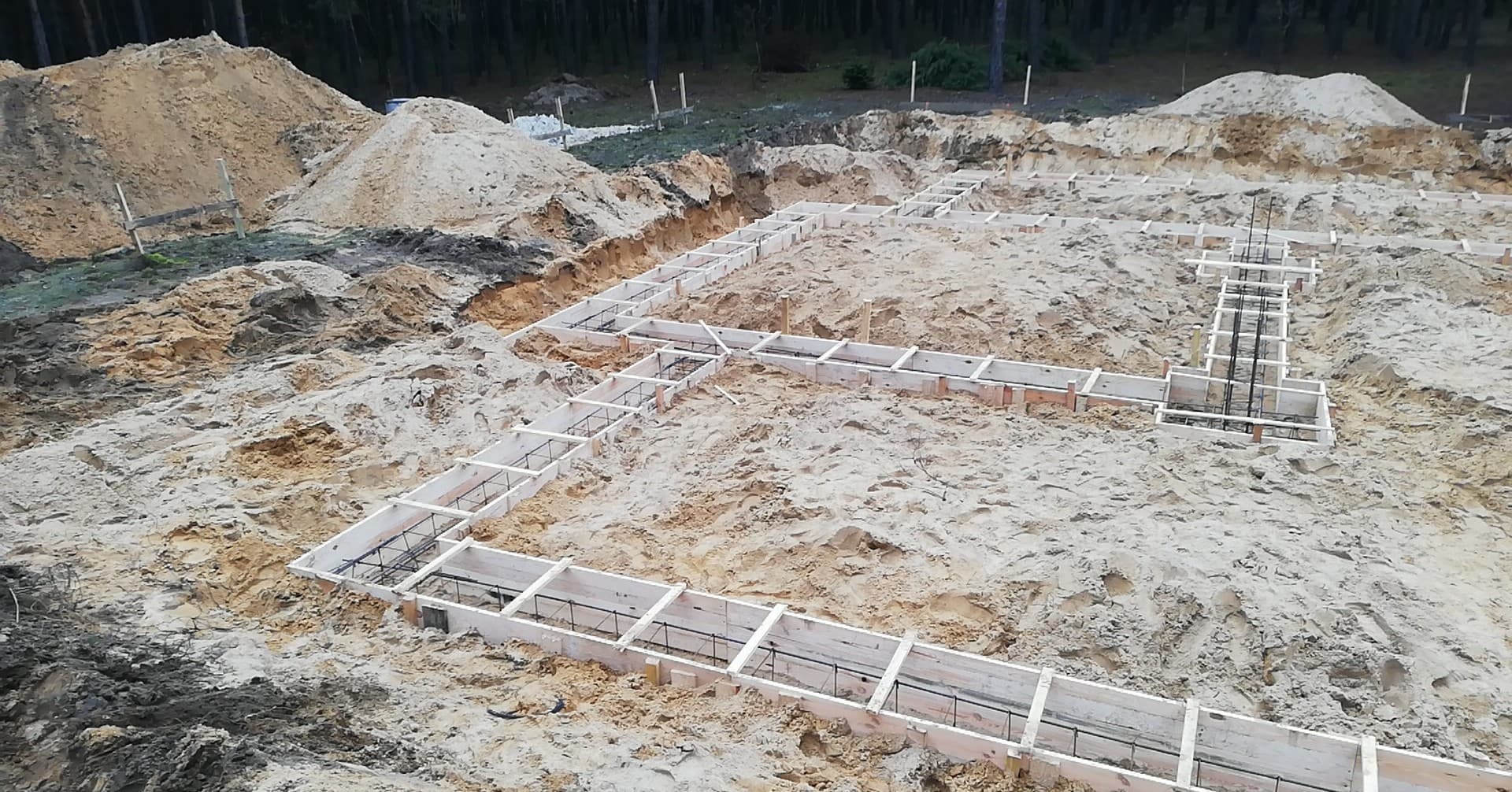 Przygotowanie fundamentów pod budowę domu: szalunek z drewnianych desek i stalowe zbrojenie w wykopie na piaszczystym terenie, widoczne pryzmy ziemi i drzewa w tle.