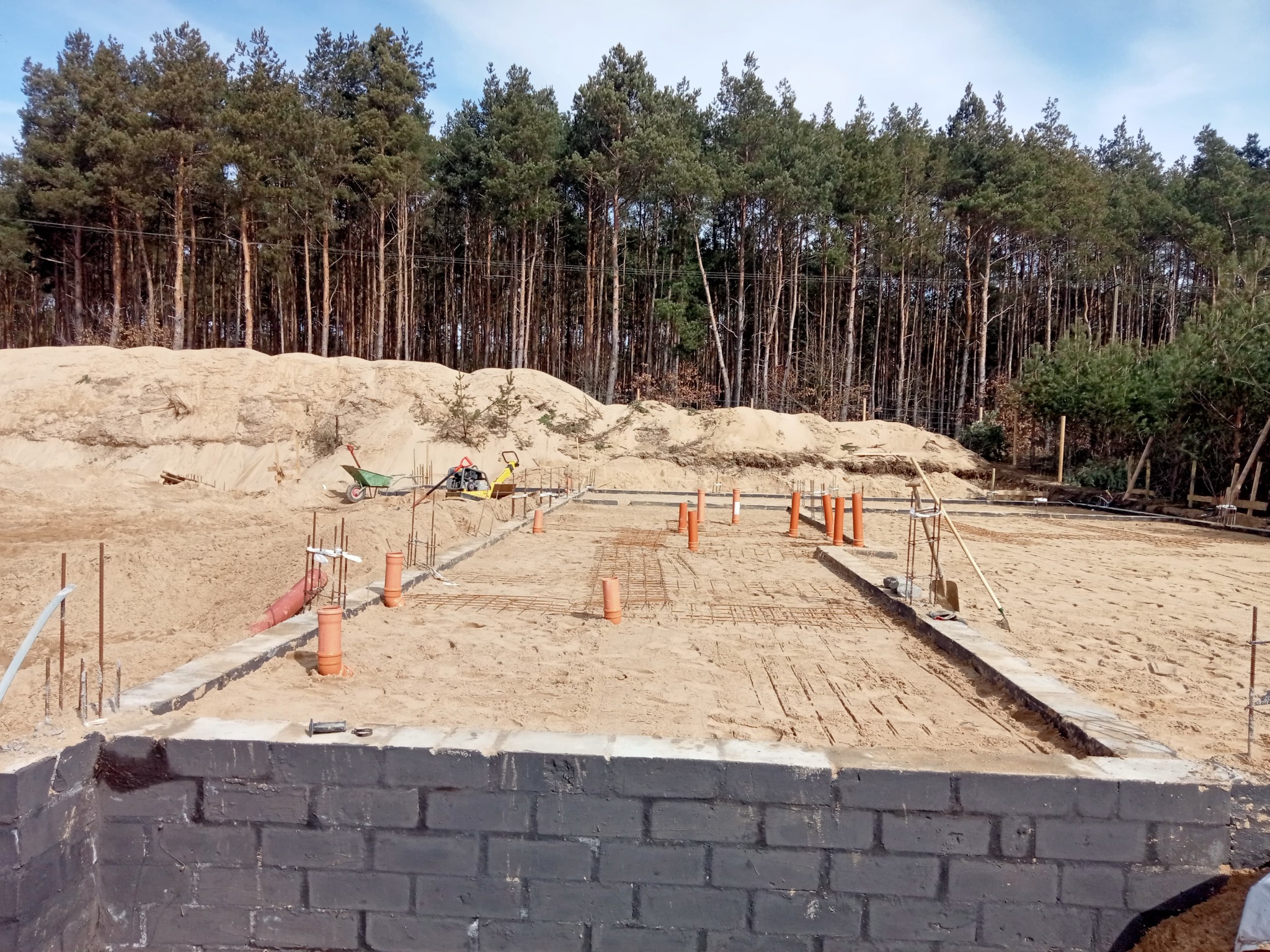 Budowa fundamentów domu jednorodzinnego z widoczną zbrojoną ławą fundamentową, pomalowanymi bloczkami betonowymi oraz pionowymi rurami kanalizacyjnymi wystającymi z piasku, w tle las i sprzęt...