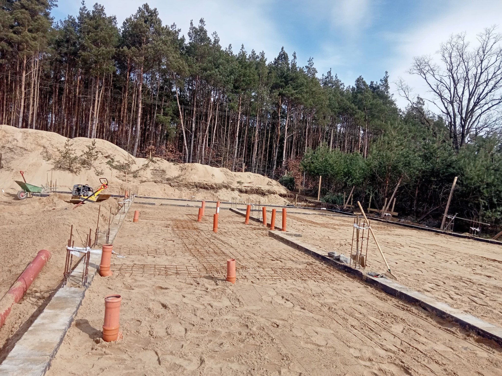 Przygotowanie terenu pod budowę domu w Toruniu: widoczne fundamenty, wystające rury kanalizacyjne i zbrojenia na piaszczystym podłożu, w tle las.