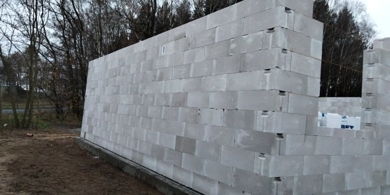 Ściana w trakcie budowy z szarych bloczków betonowych, widoczne spoiny i otwory montażowe, w tle drzewa bez liści i fragment zapakowanych materiałów budowlanych.
