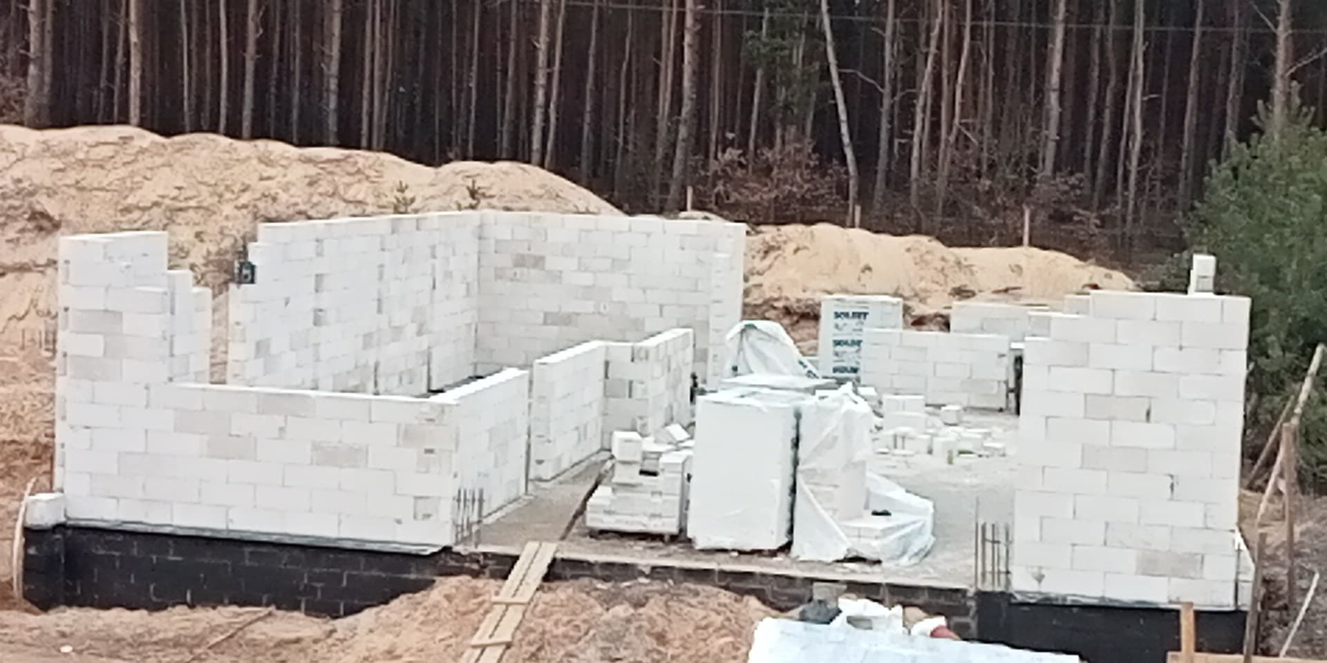 Wznoszone ściany domu z białych bloczków, widoczne fundamenty, w tle las i hałda piasku.
