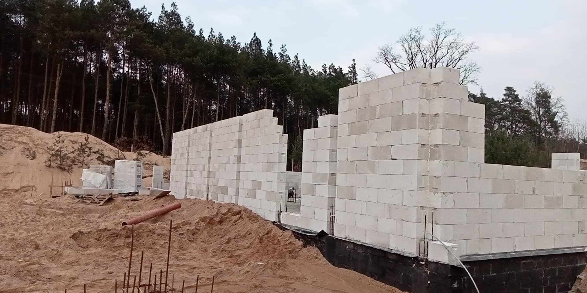 Wznoszenie ścian domu z bloczków na fundamencie, widoczny piasek i las w tle.