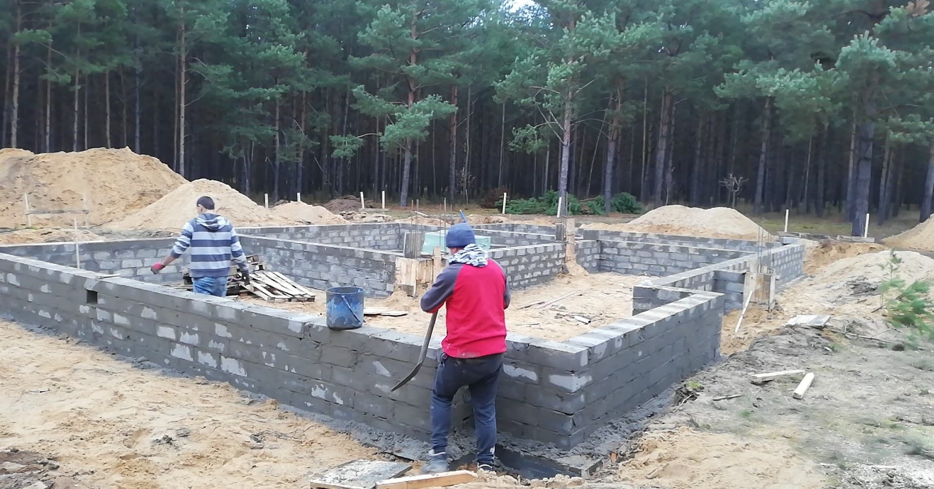 Fundamenty domu z bloczków betonowych na piaszczystym terenie, w tle las sosnowy, dwóch pracowników budowlanych przy pracy.