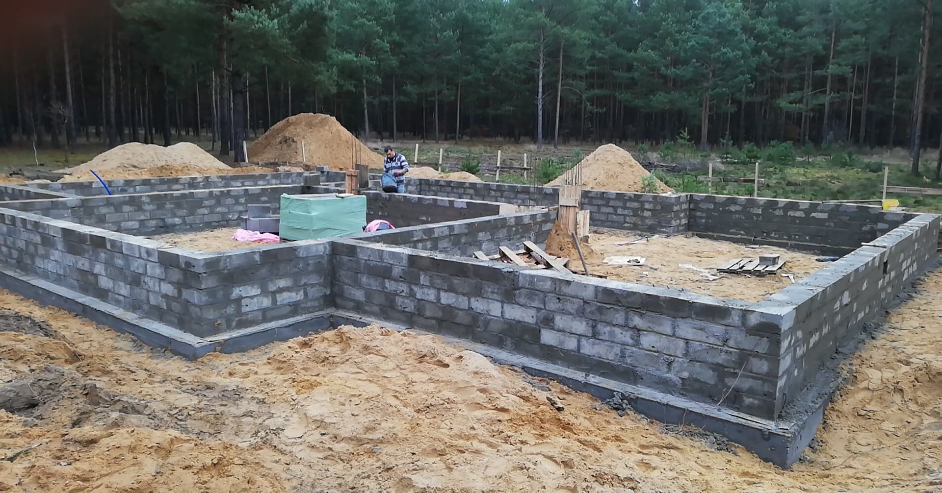 Wznoszenie fundamentów z bloczków betonowych na piaszczystym terenie, widoczny pracownik w tle oraz stosy piasku.