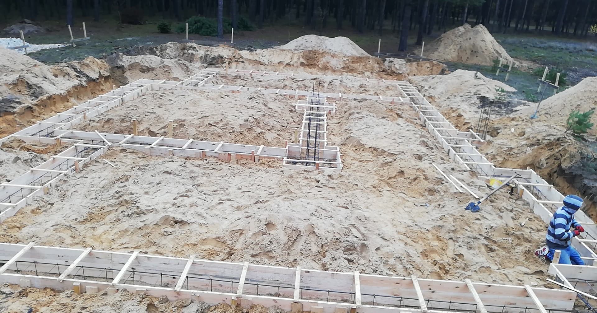 Przygotowanie fundamentów pod budowę domu murowanego: drewniany szalunek zbrojony stalą, widok z góry na wykop z piaskiem, w tle las i hałda ziemi, w rogu kadru pracownik w niebieskiej czapce...