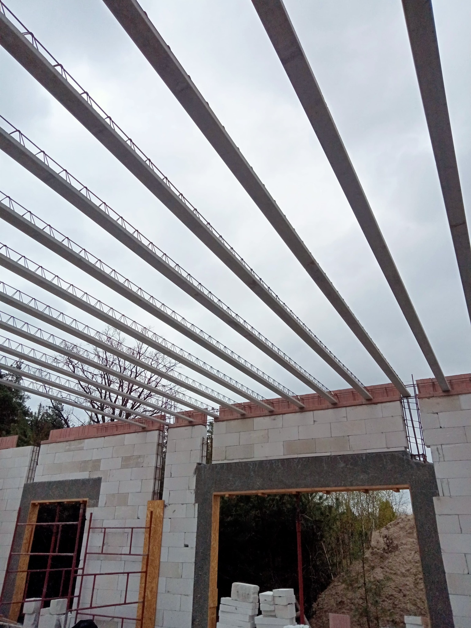 Budowa domu murowanego: widok na strop z belek i pustaków ceramicznych, ściany z betonu komórkowego, w tle drzewa i pochmurne niebo.