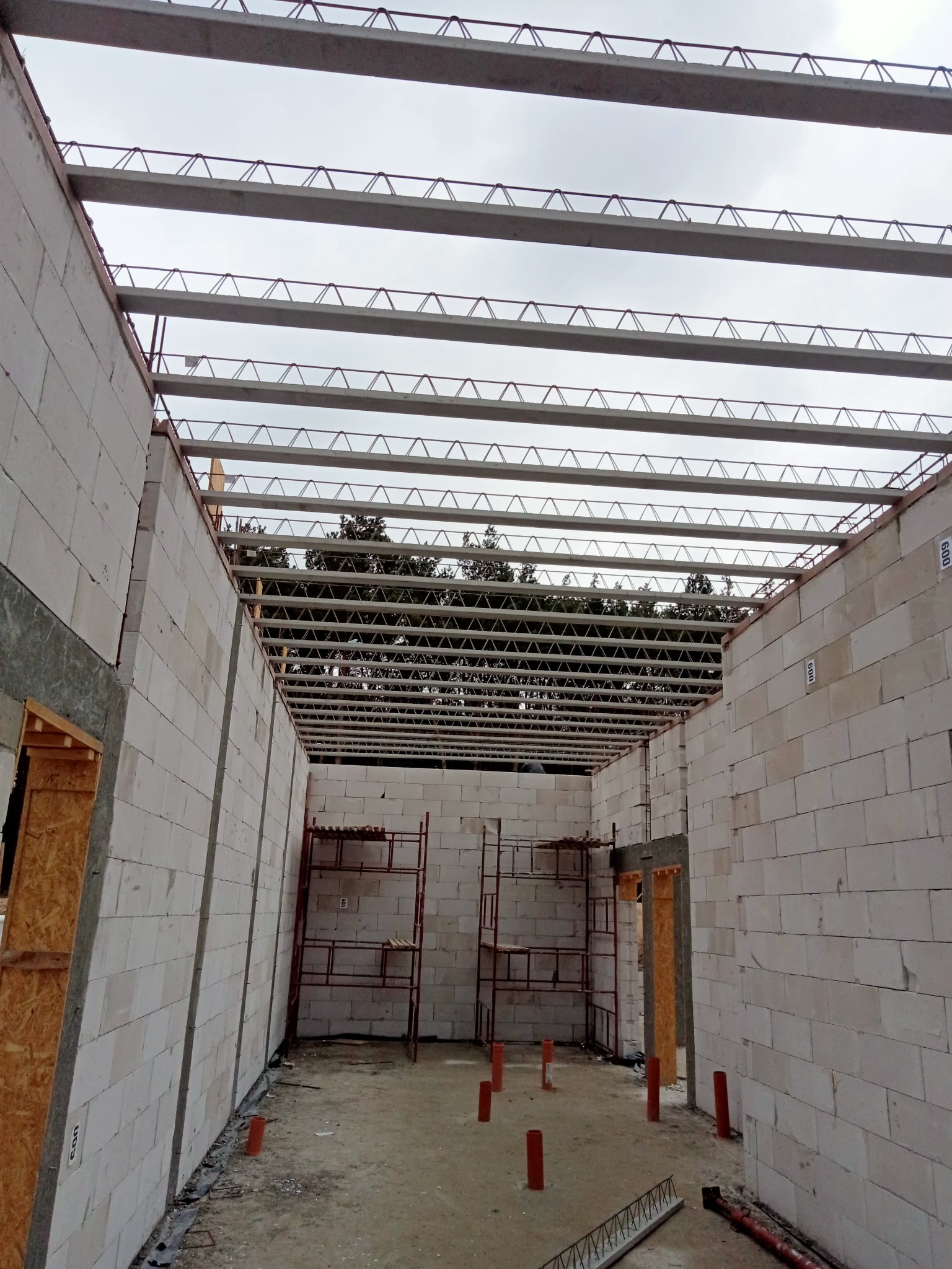 Wnętrze domu w trakcie budowy z białych bloczków, widoczne stalowe belki stropowe i rusztowanie, na zewnątrz zieleń drzew.