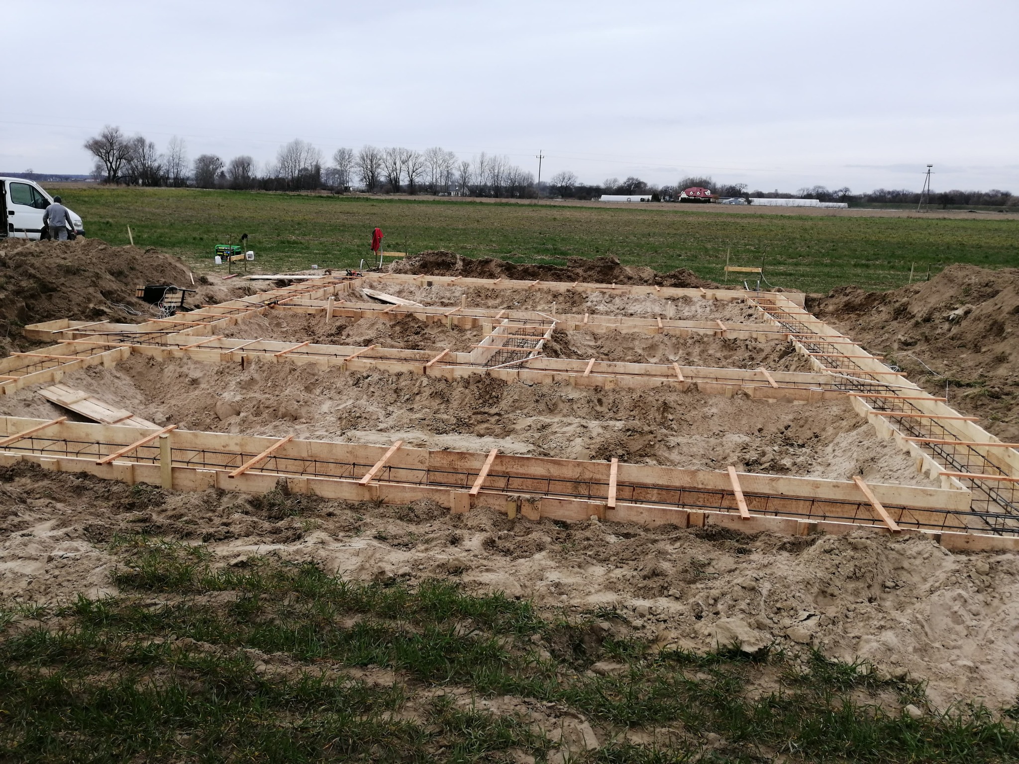 Wykonana drewniana konstrukcja szalunkowa zbrojona stalą na fundamencie pod budowę domu jednorodzinnego, widok na otwarty teren z samochodem dostawczym i pracownikiem w tle.