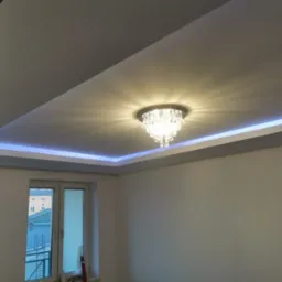 Kryształowy żyrandol zamontowany w podwieszanym suficie z oświetleniem LED, widok na okno i fragment budynku za nim.