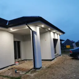 Nowoczesny dom jednorodzinny z oświetleniem zewnętrznym w podbitce, świeżo po nałożeniu jasnego tynku, widoczne ślady prac wykończeniowych na gruncie.