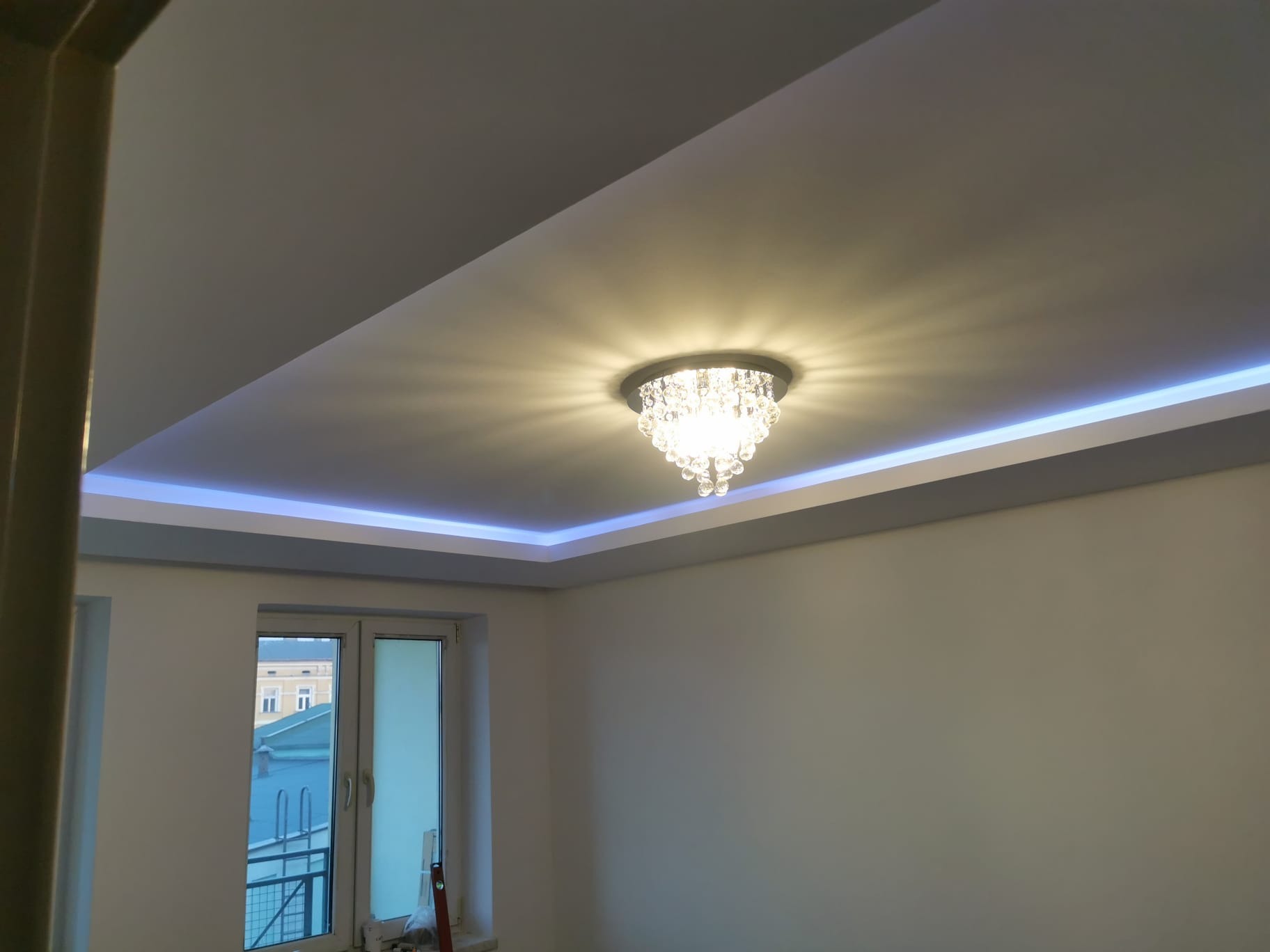 Kryształowy żyrandol zamontowany w podwieszanym suficie z oświetleniem LED, widok na okno i fragment budynku za nim.