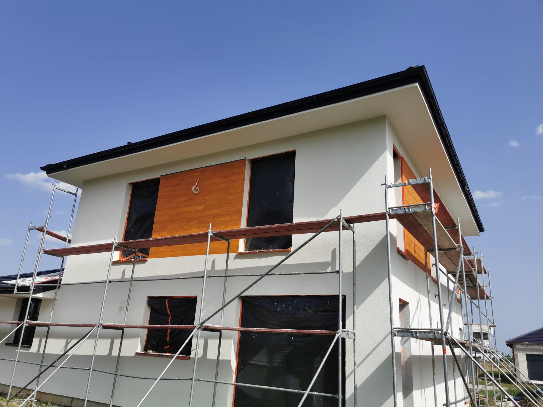 Dom w trakcie ocieplania elewacji, okna zabezpieczone folią, widoczne rusztowania, fragment drewnianej okładziny na elewacji.