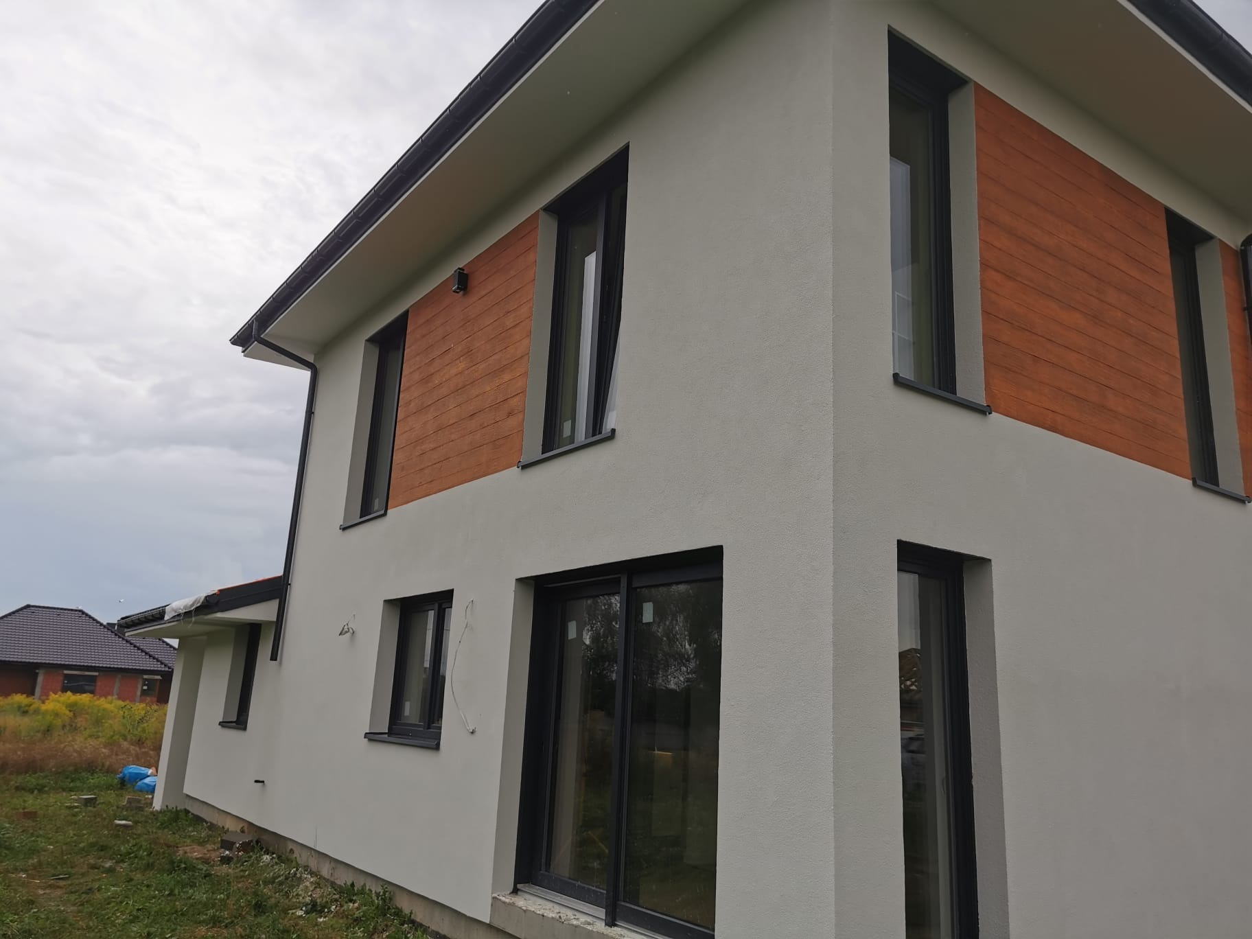 Nowoczesny dom jednorodzinny z jasnoszarą elewacją i panelami z drewna kompozytowego wokół okien, widok z zewnątrz w pochmurny dzień.