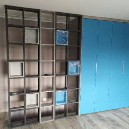 Yury Feldberh - Regał z kwadratowymi półkami o metalowej konstrukcji, niektóre z elementami jeszcze zabezpieczonymi folią, stoi obok zabudowy szafy z niebieskimi frontami i srebrnymi uchwytami na tle szarej ściany...