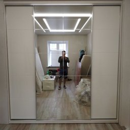 Yury Feldberh - Biała szafa przesuwna z lustrzanymi drzwiami, odbijająca wnętrze pokoju z oknem i elementami wyposażenia w trakcie montażu, widoczne narzędzia i materiały budowlane.