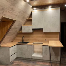 Kuchnia na poddaszu ze skośnym sufitem, szare szafki z blatem imitującym drewno, czarny zlew i bateria, drewniana boazeria na ścianie, widoczne schodkowe szafki pod sufitem.