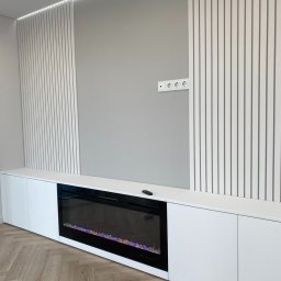 Yury Feldberh - Nowoczesna biała szafka RTV z elektrycznym kominkiem, dekoracyjnymi lamelami ściennymi i ukrytym oświetleniem LED w górnej części. Na blacie szafki leży czarny pilot.