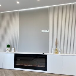 Yury Feldberh - Nowoczesna tumba pod telewizor z elektrycznym kominkiem, białe matowe fronty, pionowe panele dekoracyjne na ścianie, drewniana podłoga w jodełkę.