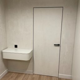 Yury Feldberh - Nowoczesna, minimalistyczna aranżacja wnętrza z ukrytymi drzwiami z czarną obwódką i wiszącą szafką nocną z zaokrąglonym frontem, na tle ściany o fakturze betonu.