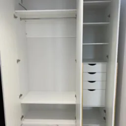 Wnętrze białej, otwartej szafy z półkami, drążkiem na ubrania i czterema szufladami, z widocznymi zawiasami i marmurową podłogą w tle.