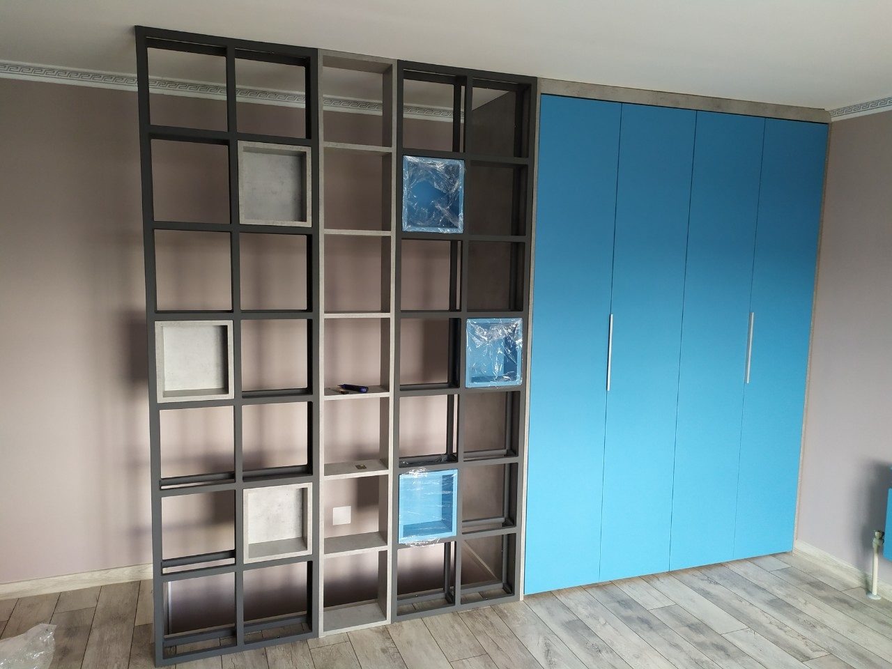 Regał z kwadratowymi półkami o metalowej konstrukcji, niektóre z elementami jeszcze zabezpieczonymi folią, stoi obok zabudowy szafy z niebieskimi frontami i srebrnymi uchwytami na tle szarej ściany...